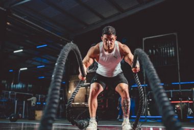 Focused man in white tank top performing battle rope exercise in a modern gym.